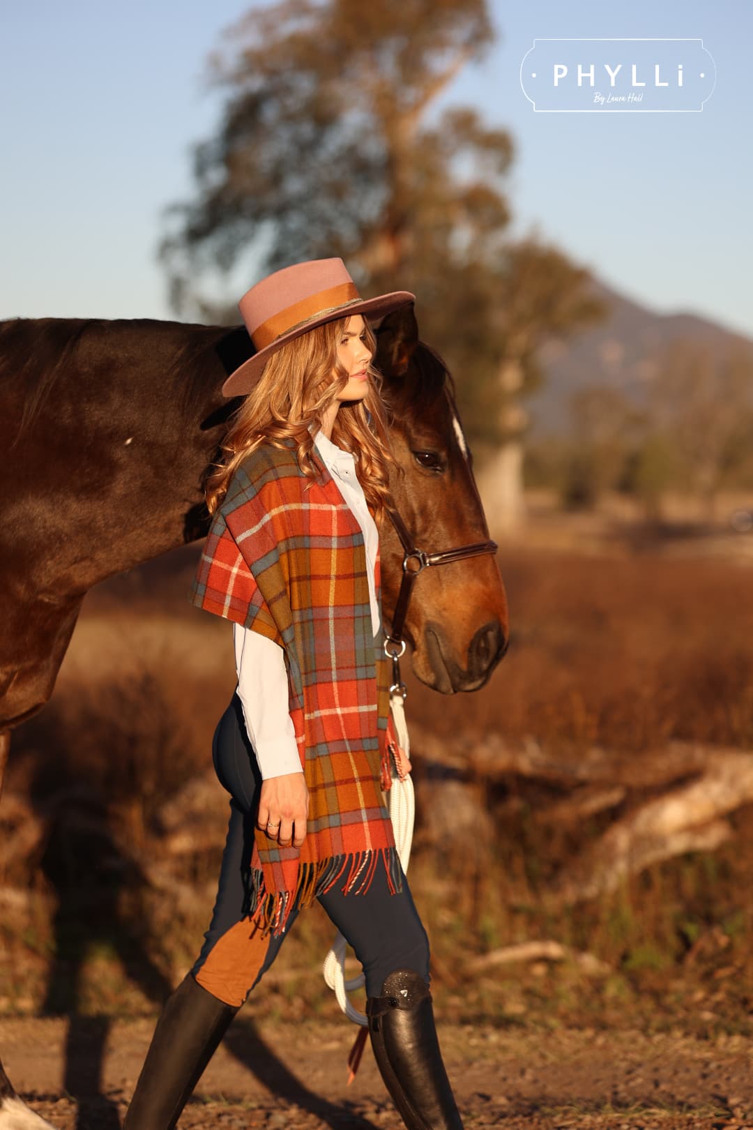 Full body image of woman wearing The Blush felt hat and plaid wrap walking with horse in countryside.