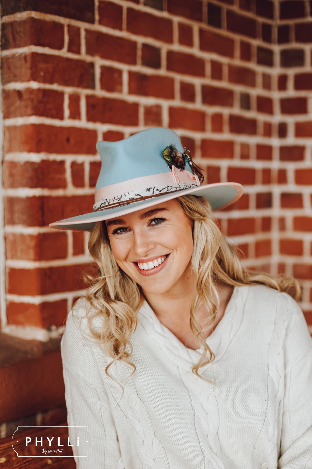 Blonde model wearing The Cyan felt hat by PHYLLi Designs with sherbet pink grosgrain band and silver chunky hat pin against brick wall background.