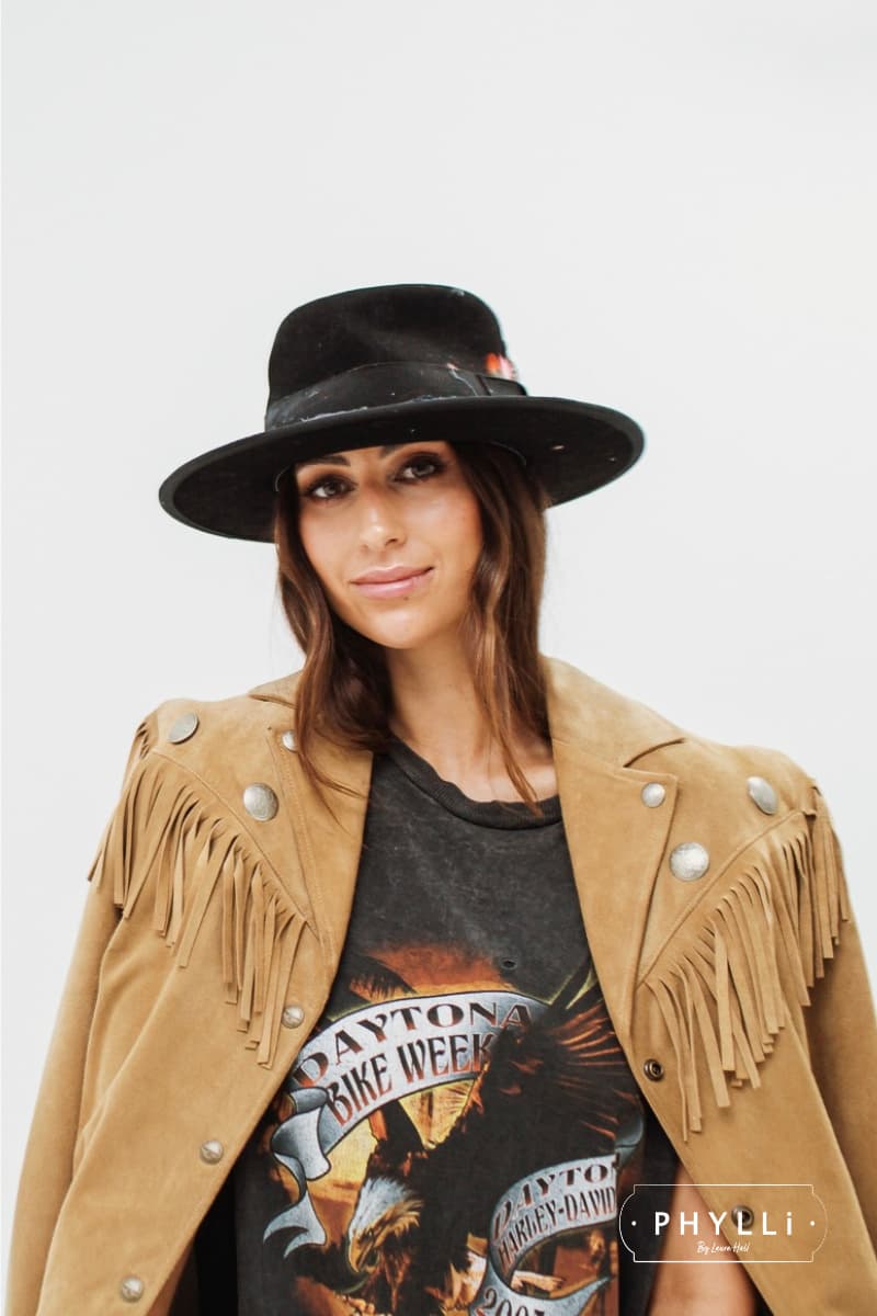 Woman wearing a black felt hat by PHYLLi Designs styled with a tan fringe jacket and graphic t-shirt.