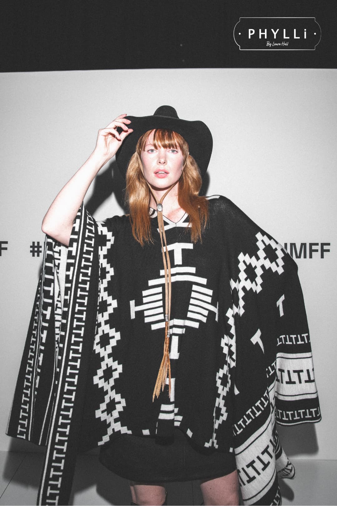 Model wearing The PHYLLi dark navy felt hat by PHYLLi Designs with Buffalo Girl Rugged Desert tie back band at a fashion event backdrop.