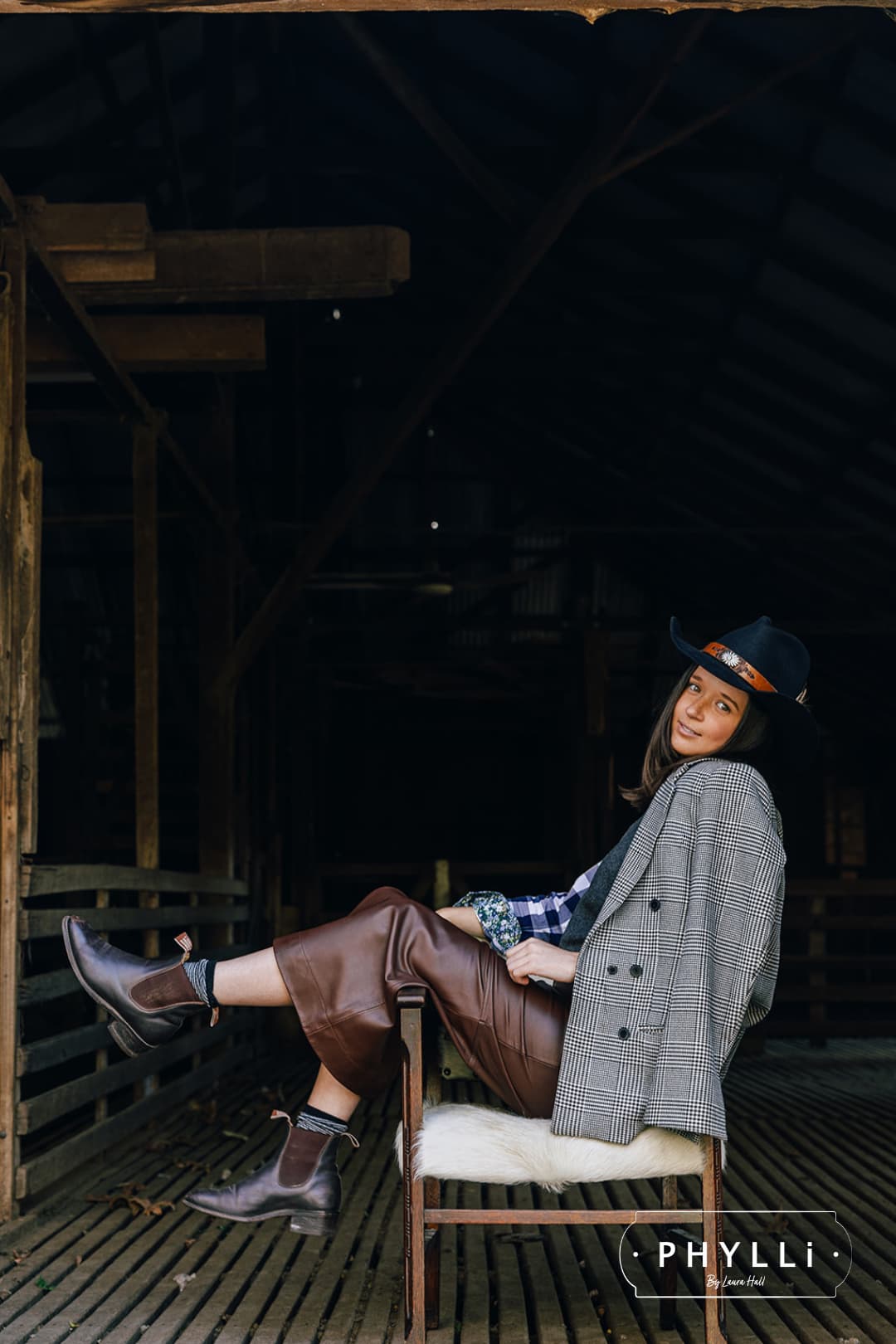 Model seated wearing The PHYLLi dark navy felt hat by PHYLLi Designs with feather trim and leather band in a barn interior.