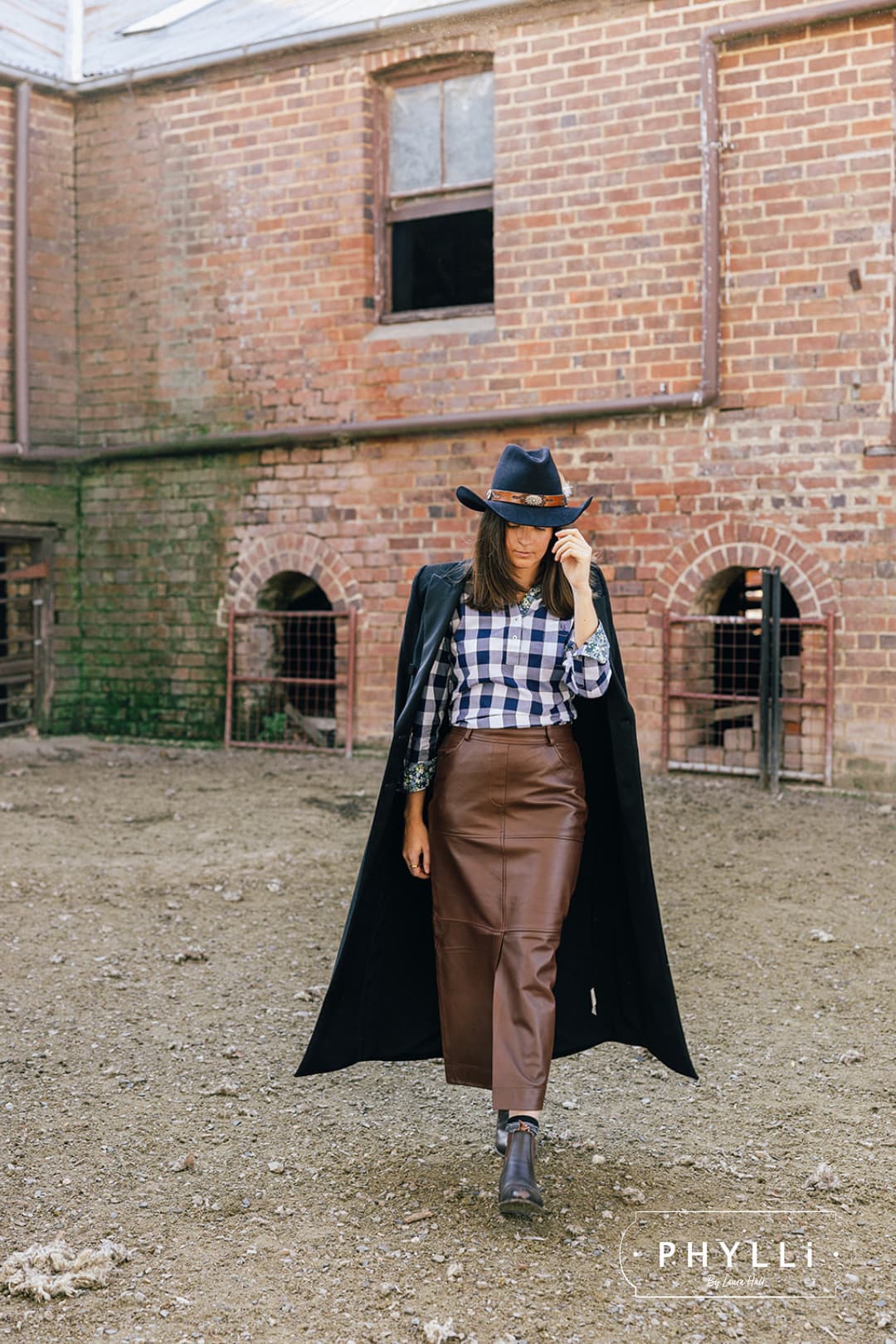 Model wearing The PHYLLi dark navy felt hat by PHYLLi Designs with feather trim in an outdoor rural heritage setting.