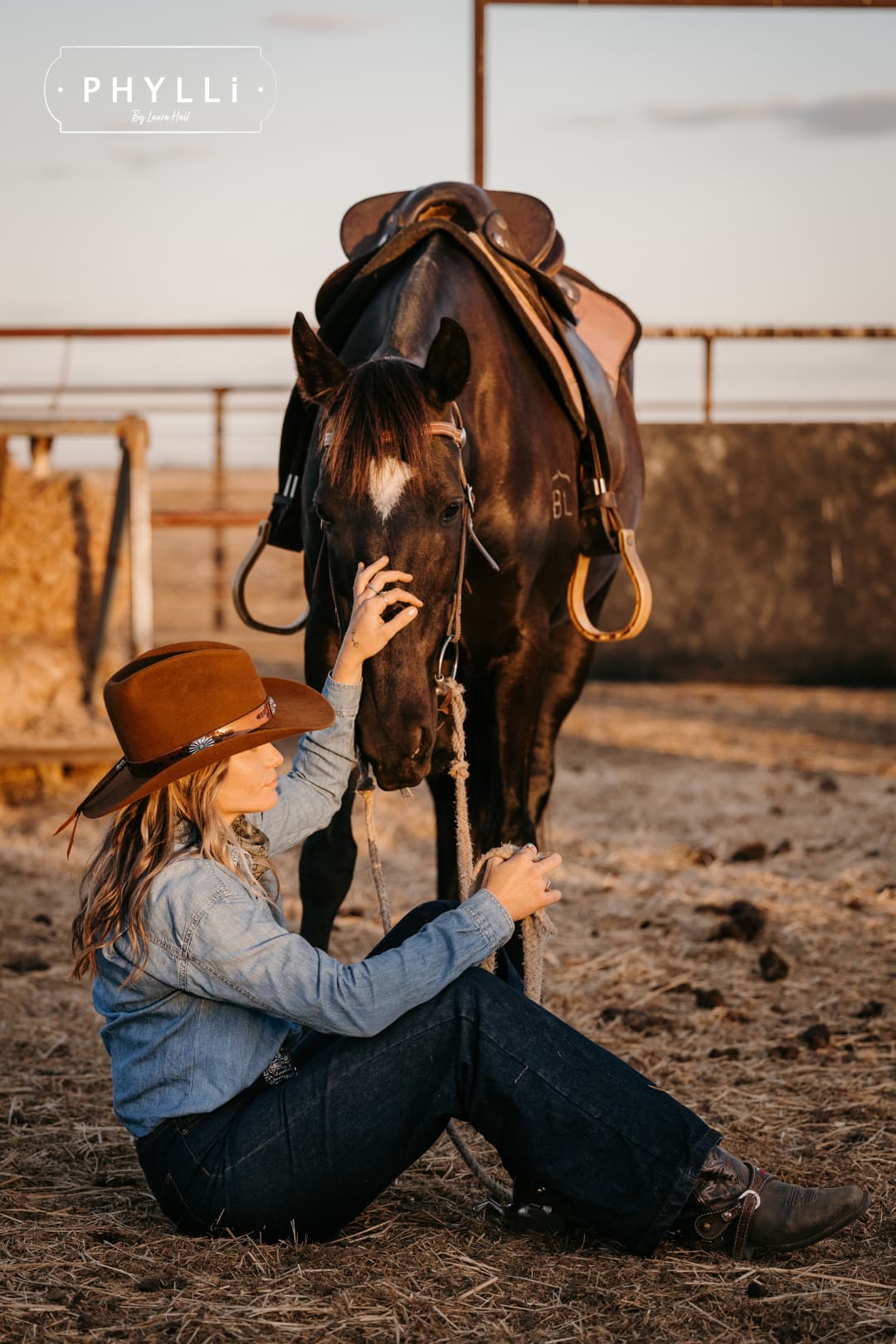 Woman wearing the Caramel cowboy hat The PHYLLi Caramel by PHYLLi Designs with Buffalo Girl Western floral tie back hat band sitting beside a saddled horse in a rural paddock setting.