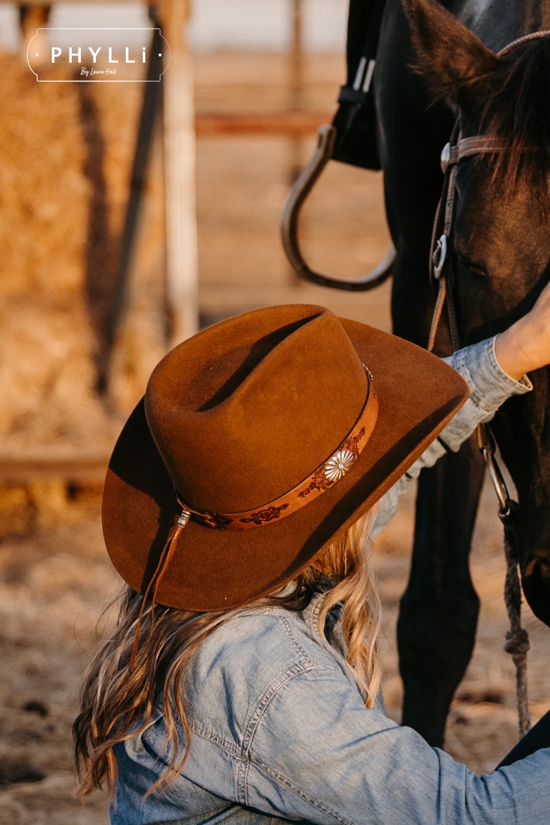 Woman wearing the Caramel cowboy hat The PHYLLi Caramel by PHYLLi Designs with Buffalo Girl Western floral tie back hat band while sitting beside a horse in a rustic stockyard.