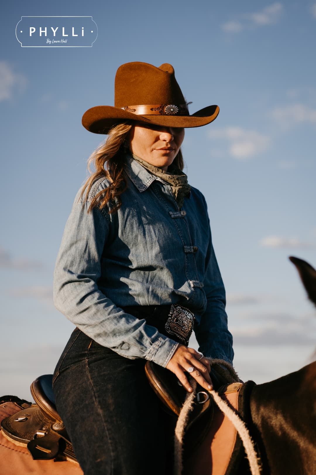 Cowgirl wearing the Caramel cowboy hat The PHYLLi Caramel by PHYLLi Designs with Buffalo Girl Western floral tie back hat band while seated beside a horse against a blue sky.