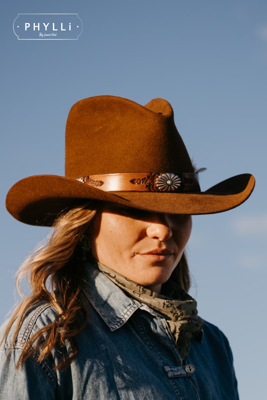 Close up cowgirl wearing the Caramel cowboy hat The PHYLLi Caramel by PHYLLi Designs with Buffalo Girl Western floral tie back hat band in an open rural paddock at sunset.