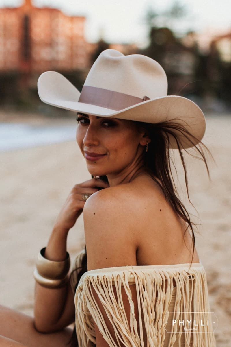 Woman wearing The Plains beige felt cowboy hat by PHYLLi Designs with plaster grosgrain double bow band at beach location.