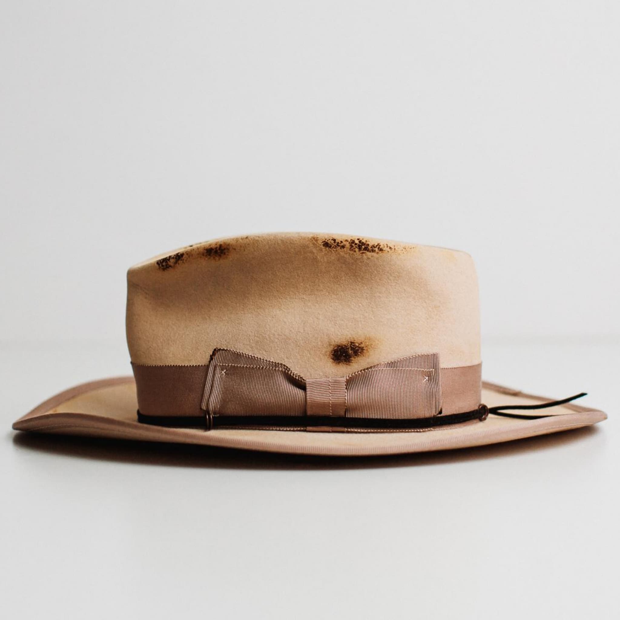 Left side view of Ringleader beige distressed felt hat by PHYLLi Designs with plaster grosgrain band, plaster brim trim and black suede string with bronze O rings.
