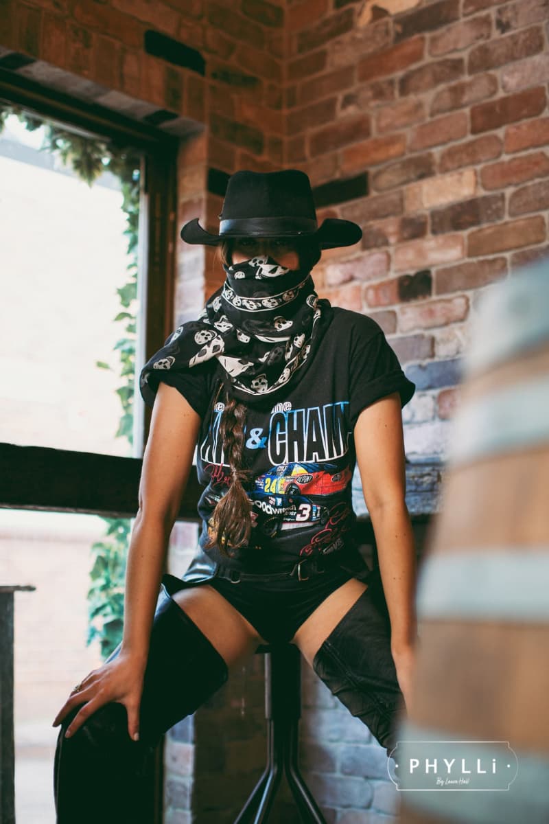Person wearing a black felt cowboy hat and bandana sitting against a brick wall.