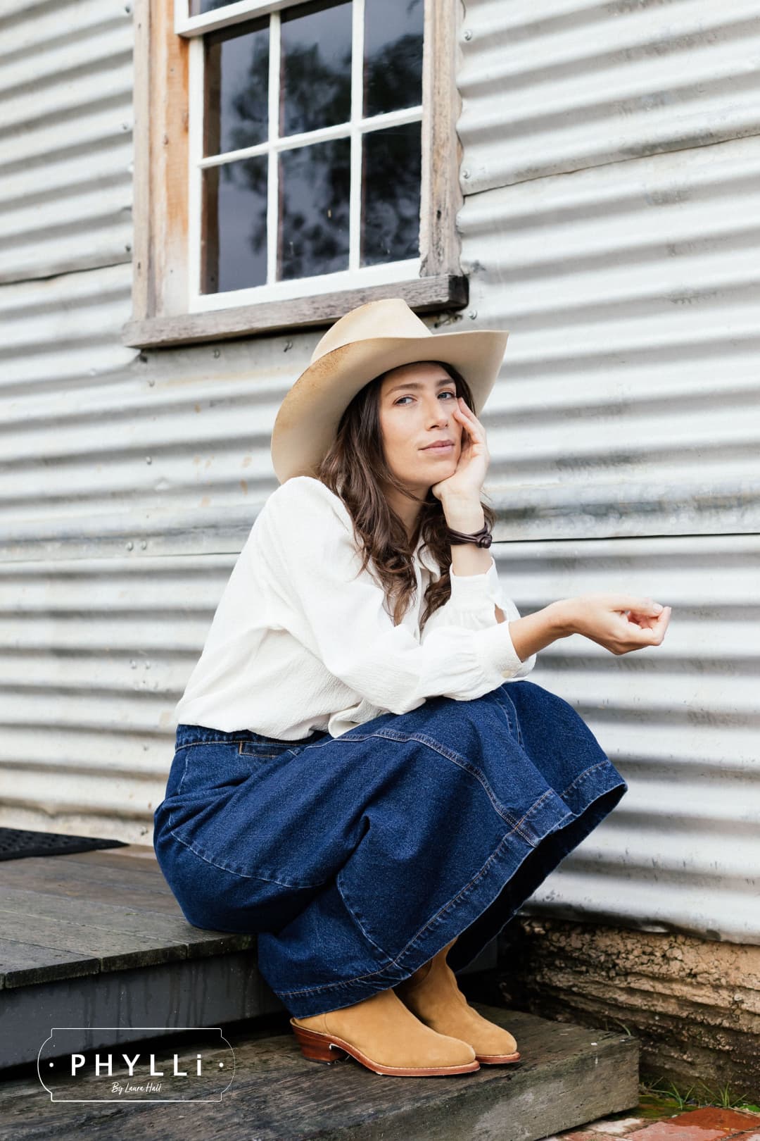 Woman seated outside farmhouse wearing The Wheeler premium felt hat in distressed Beige.