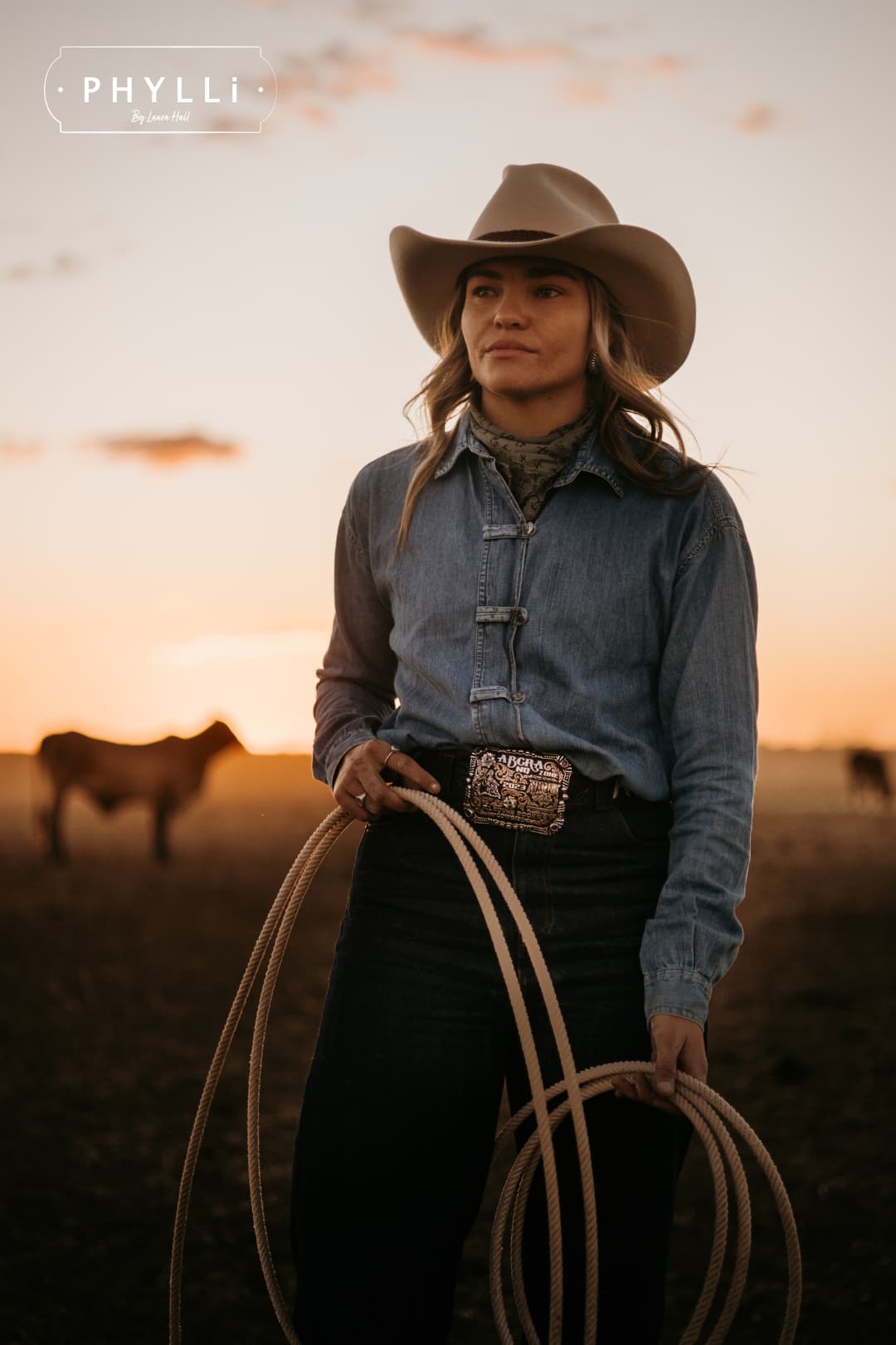 Model wearing the beige cowboy hat Wheeler Beige by PHYLLi Designs with chocolate leather hat band while holding a stockyard lariat at sunset in an open paddock.