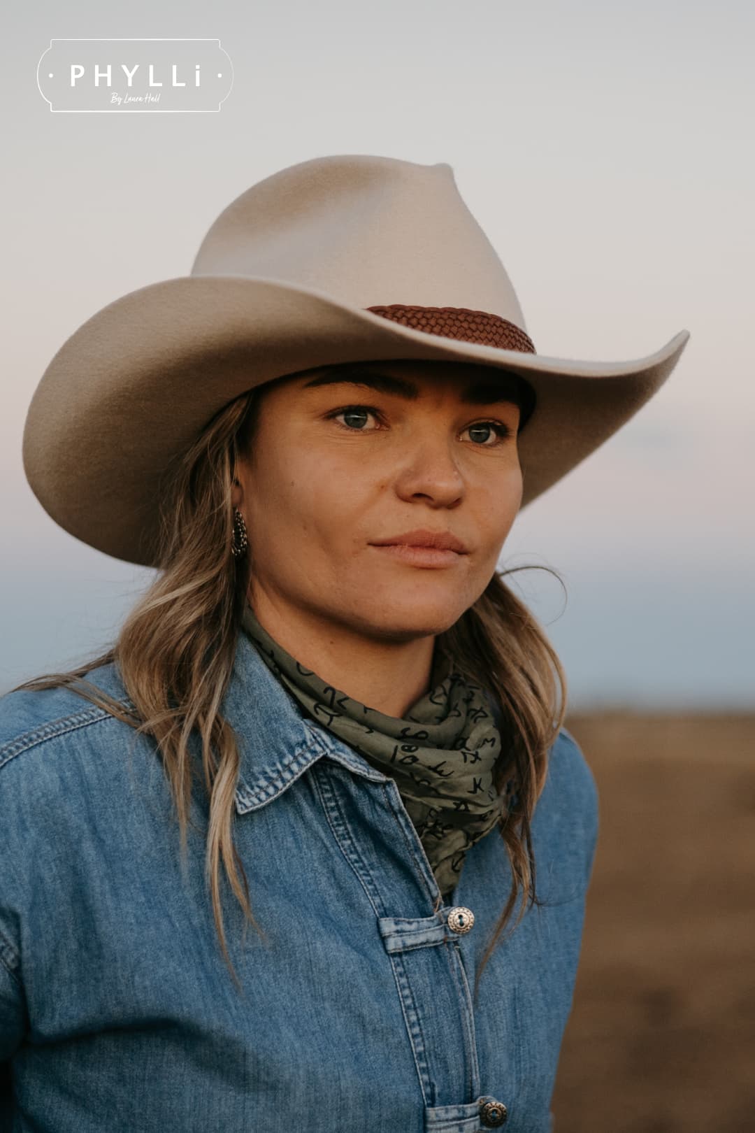 Close up model wearing the beige cowboy hat Wheeler Beige by PHYLLi Designs with 12 strand chocolate leather hat band in an open rural landscape.