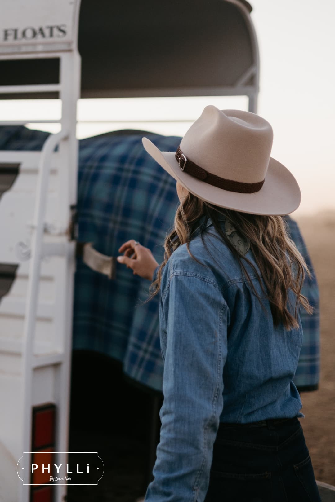 Model wearing the beige cowboy hat Wheeler Beige by PHYLLi Designs with chocolate leather hat band viewed from behind beside a horse float.