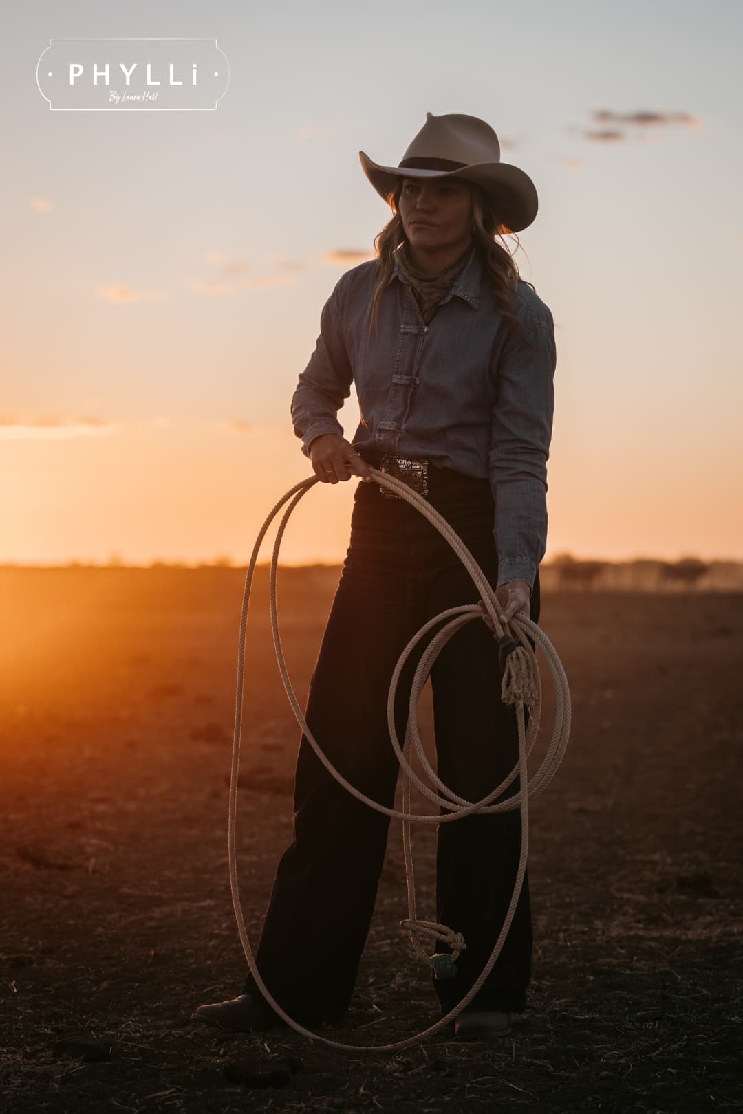 Model wearing the beige cowboy hat Wheeler Beige by PHYLLi Designs with chocolate leather hat band holding a coiled lariat in a sunlit paddock at sunset.