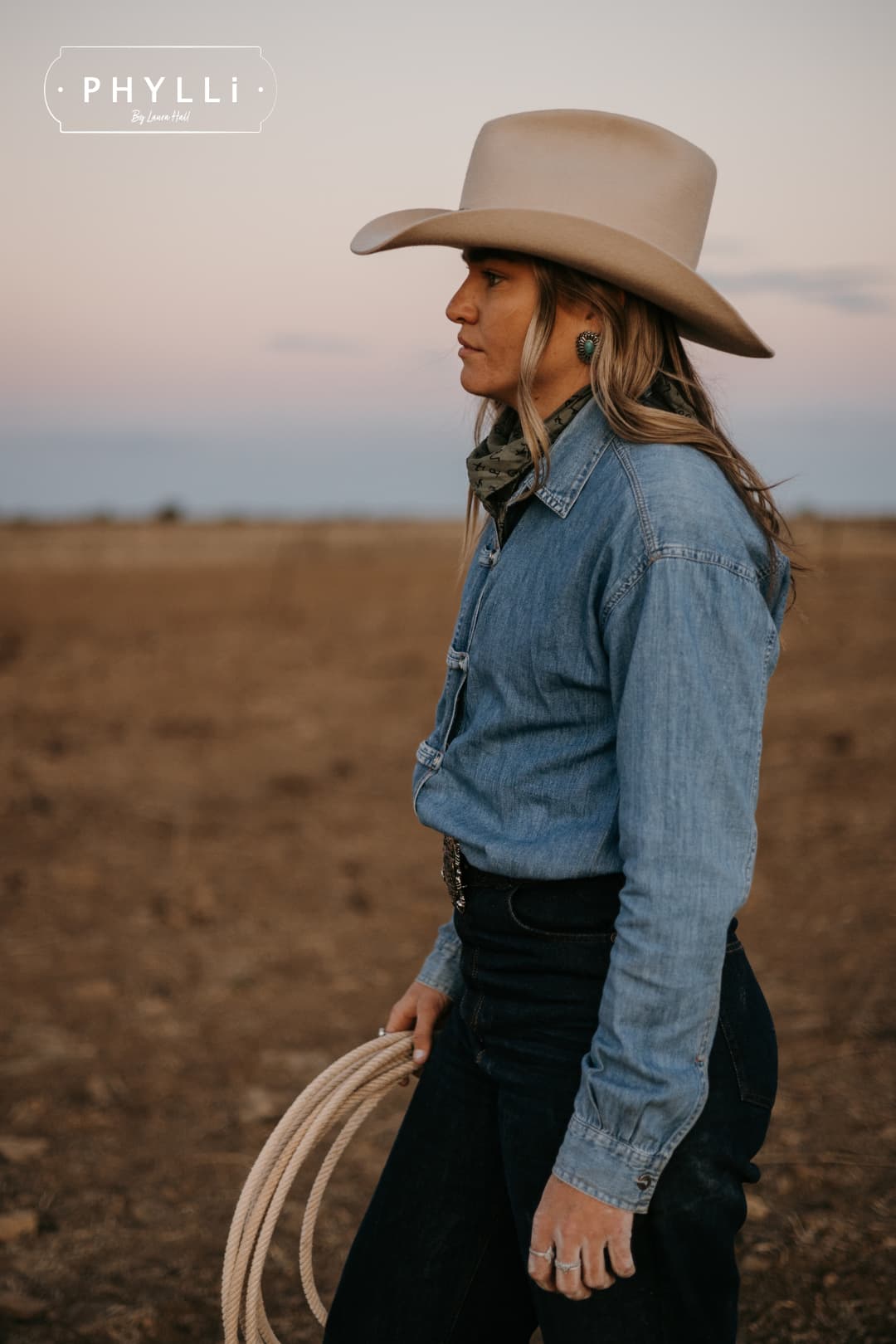 Model wearing the beige cowboy hat Wheeler Beige by PHYLLi Designs with chocolate leather hat band holding a coiled stockyard lariat in an open rural paddock.