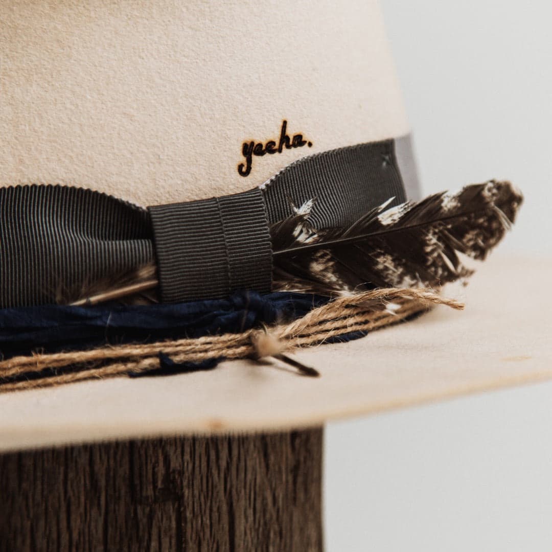 Close up detail of the Wrangler beige distressed felt hat by PHYLLi Designs showing burnt branded yeeha text above the steel grey grosgrain bow with feather.