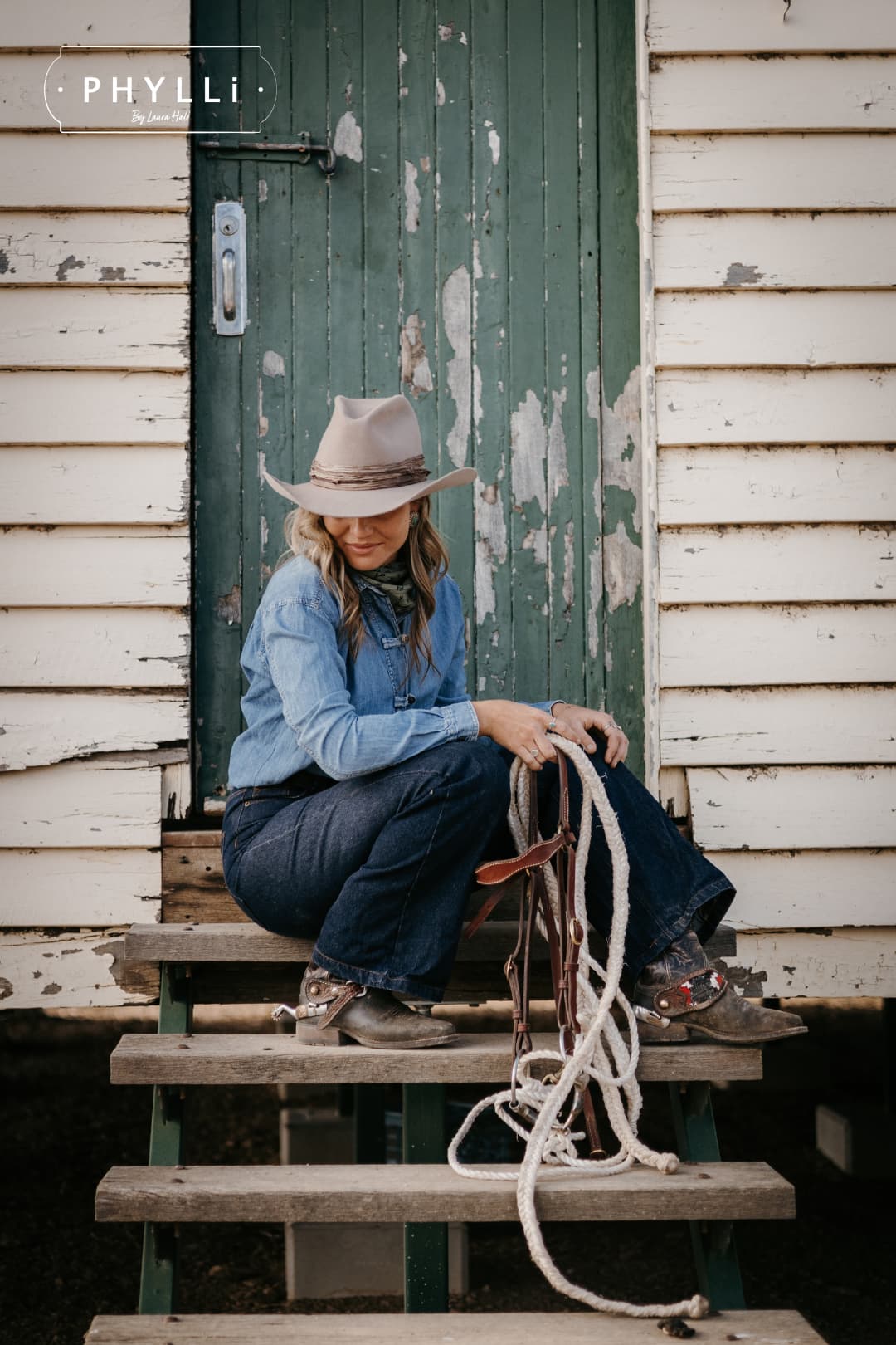 Woman wearing the Sahara cowboy hat Wrapped Wheeler Sahara by PHYLLi Designs with tonal tie-dye sari silk wrap sitting on wooden steps holding a stockman rope and leather bridle outside a weathered rural shed.