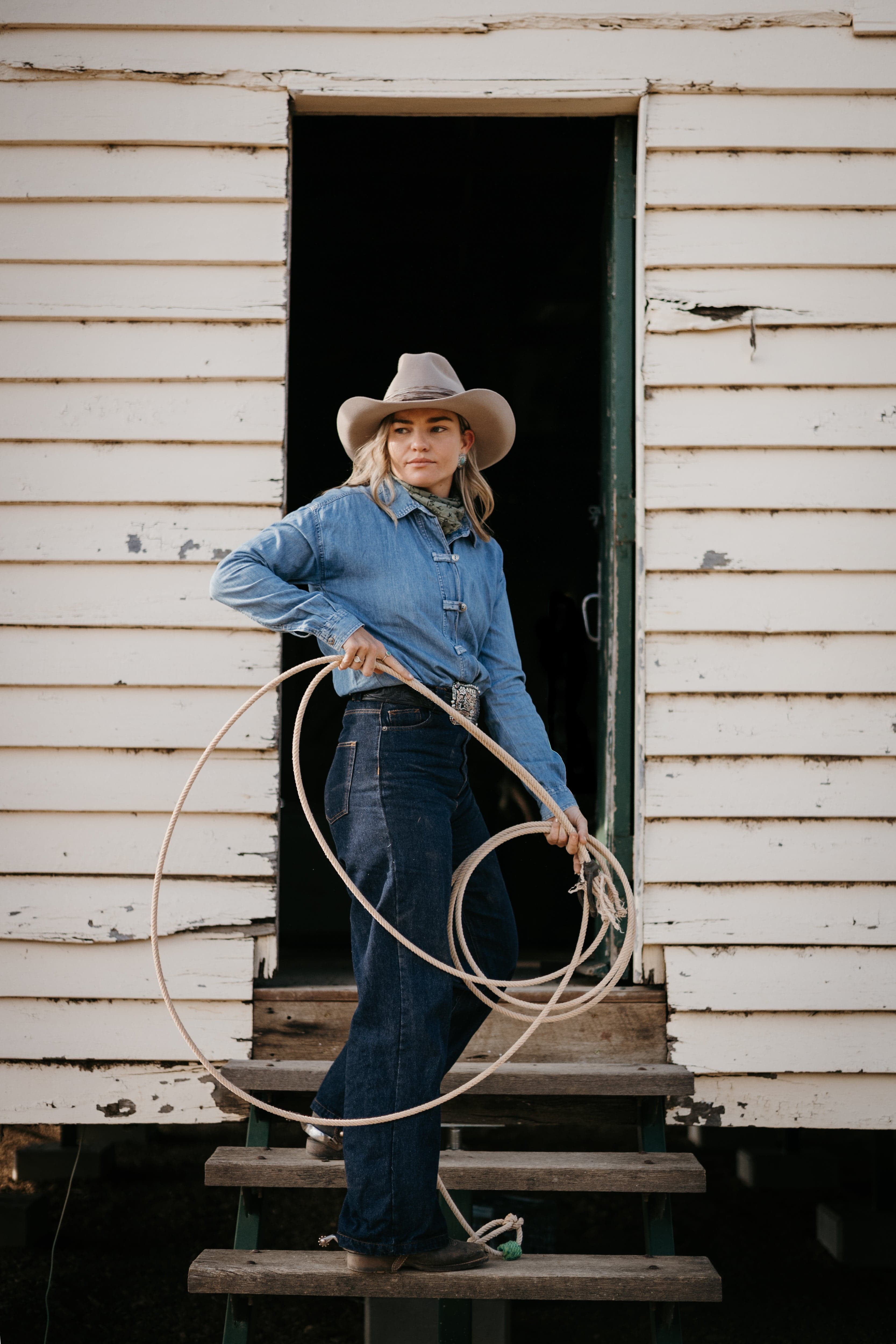 Woman wearing the Sahara cowboy hat Wrapped Wheeler Sahara by PHYLLi Designs with tonal tie-dye sari silk wrap standing in a rustic shed doorway holding a stockman rope.