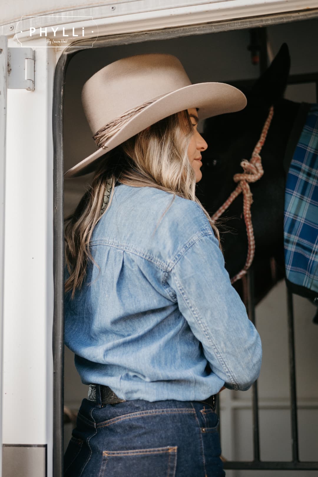 Woman wearing the Sahara cowboy hat Wrapped Wheeler Sahara by PHYLLi Designs with tonal tie-dye sari silk wrap standing in a horse float doorway beside a rugged horse.