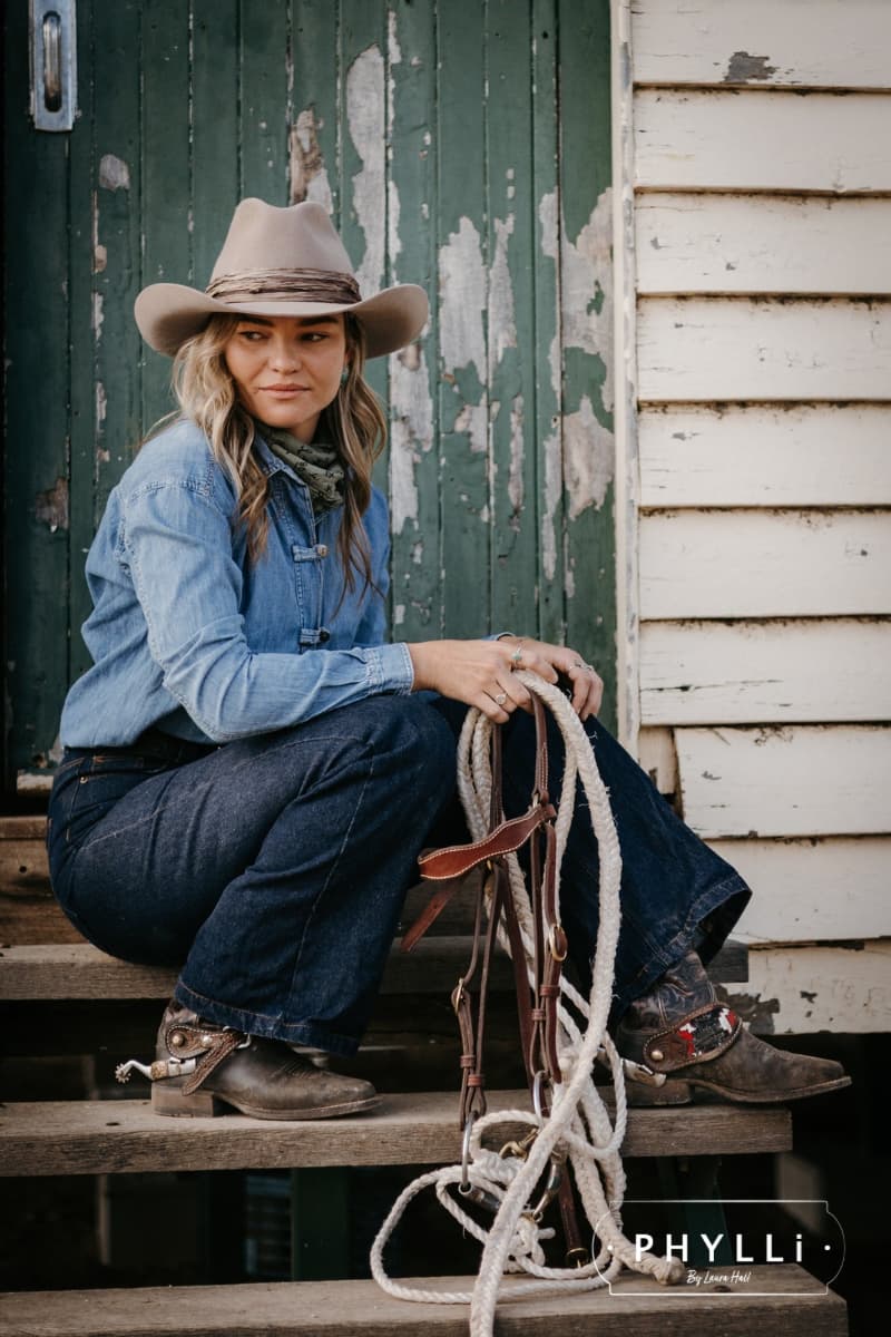 Woman wearing the Sahara cowboy hat Wrapped Wheeler Sahara by PHYLLi Designs with tonal tie-dye sari silk wrap sitting on shed steps holding rope tack and leather bridle.
